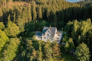 A large, two-story house surrounded by dense forest.