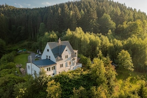 A two-story house with a sloped roof, surrounded by dense forest.