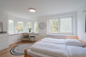 A bedroom with a bed, a round mirror, a floor lamp, a chair, and a view of trees through large windows.