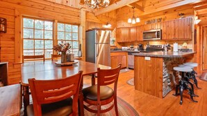 A wooden cabin interior with a large dining table, chairs, a kitchen with wooden cabinets, a refrigerator, a microwave, and a stone fireplace. - Family Cabin on 20 Private Acres • Game Room • Near Berkeley Springs (Hancock)
