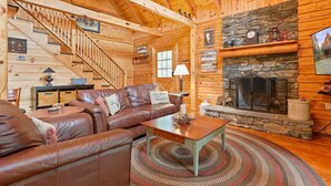 A cozy log cabin interior with a stone fireplace, leather sofa, and a staircase. - Family Cabin on 20 Private Acres • Game Room • Near Berkeley Springs (Hancock)
