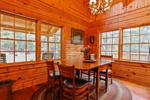 A wooden cabin interior with a dining table, chairs, and large windows offering a view of the outdoors. - Family Cabin on 20 Private Acres • Game Room • Near Berkeley Springs (Hancock)