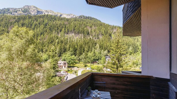 Un balcon avec une balustrade en bois, une table avec des tasses et une plante, et une vue sur une vallée boisée parsemée de maisons.
