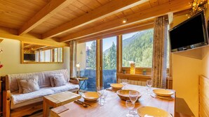 Une pièce chaleureuse avec un plafond en bois, un canapé, une table à manger dressée avec des assiettes et des verres, et un téléviseur fixé au mur.