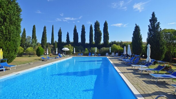A rectangular swimming pool with blue water, surrounded by lounge chairs and umbrellas, with tall trees in the background. - Casa Guna, Asciano, Italy (Asciano)