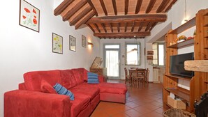 A living room with a red sofa, wooden ceiling beams, a television on a wooden stand, and framed artwork on the walls. - Casa Guna, Asciano, Italy (Asciano)