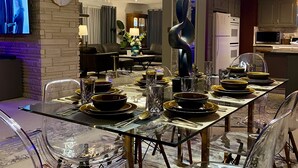 Un área de comedor moderna con una mesa y sillas de vidrio, una lámpara decorativa y una cocina al fondo.