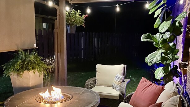 Un patio al aire libre con un brasero redondo, mobiliario de mimbre e iluminación con luces colgantes.