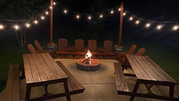 Un patio al aire libre con mesas y sillas de madera, un brasero y luces colgantes.