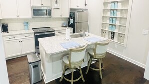 Una cucina moderna con mobili bianchi, un'isola centrale con sedie, forno a microonde, piano cottura, frigorifero e scaffale per vini.