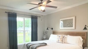 Una camera da letto con un letto grande, una ventola a soffitto, una finestra con tende, una lampada da comodino e un quadro appeso al muro.