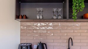 Una cocina con salpicadero de azulejos, un lavamanos de acero inoxidable, un tostador negro, una tetera y una estantería con copas de vino y una planta.
