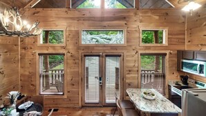 Interior de una cabaña de madera con comedor, cocina y ventanas amplias.