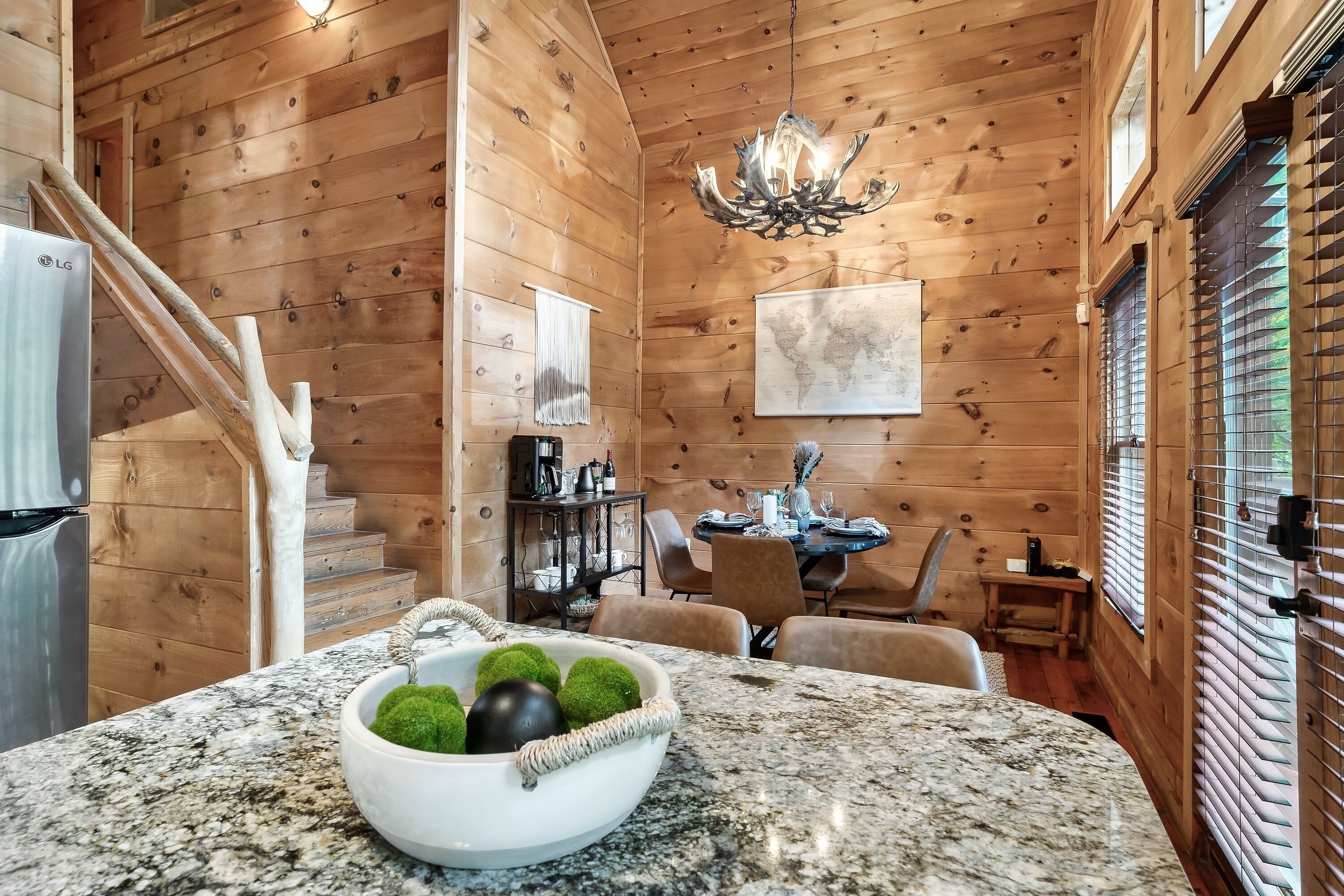 A wooden cabin interior with a kitchen area featuring a granite countertop, a bowl with green and black spherical objects, a chandelier, and a dining table with chairs.