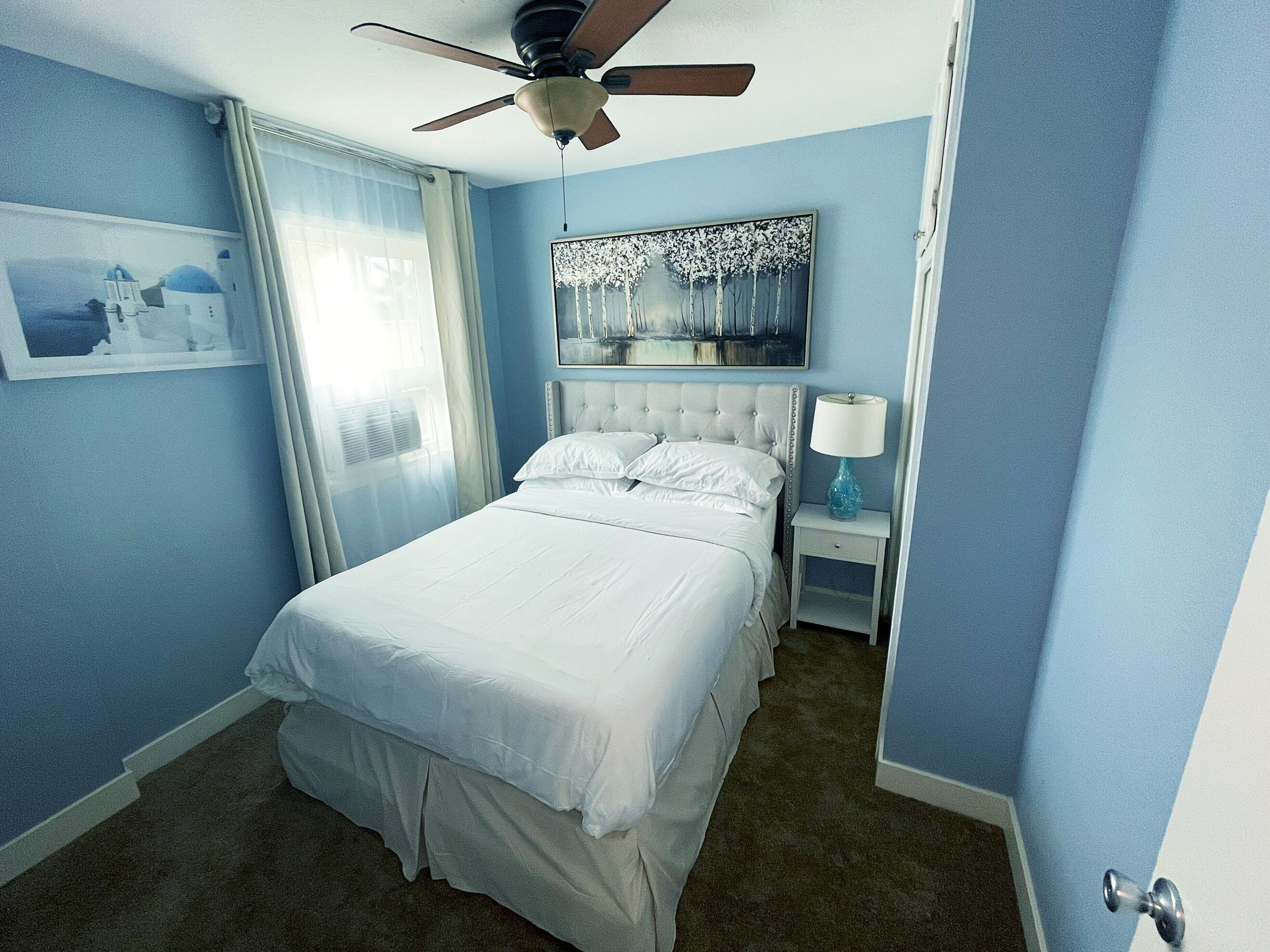 Une chambre à coucher avec un lit, un ventilateur de plafond, une table de chevet avec une lampe et un tableau au mur.