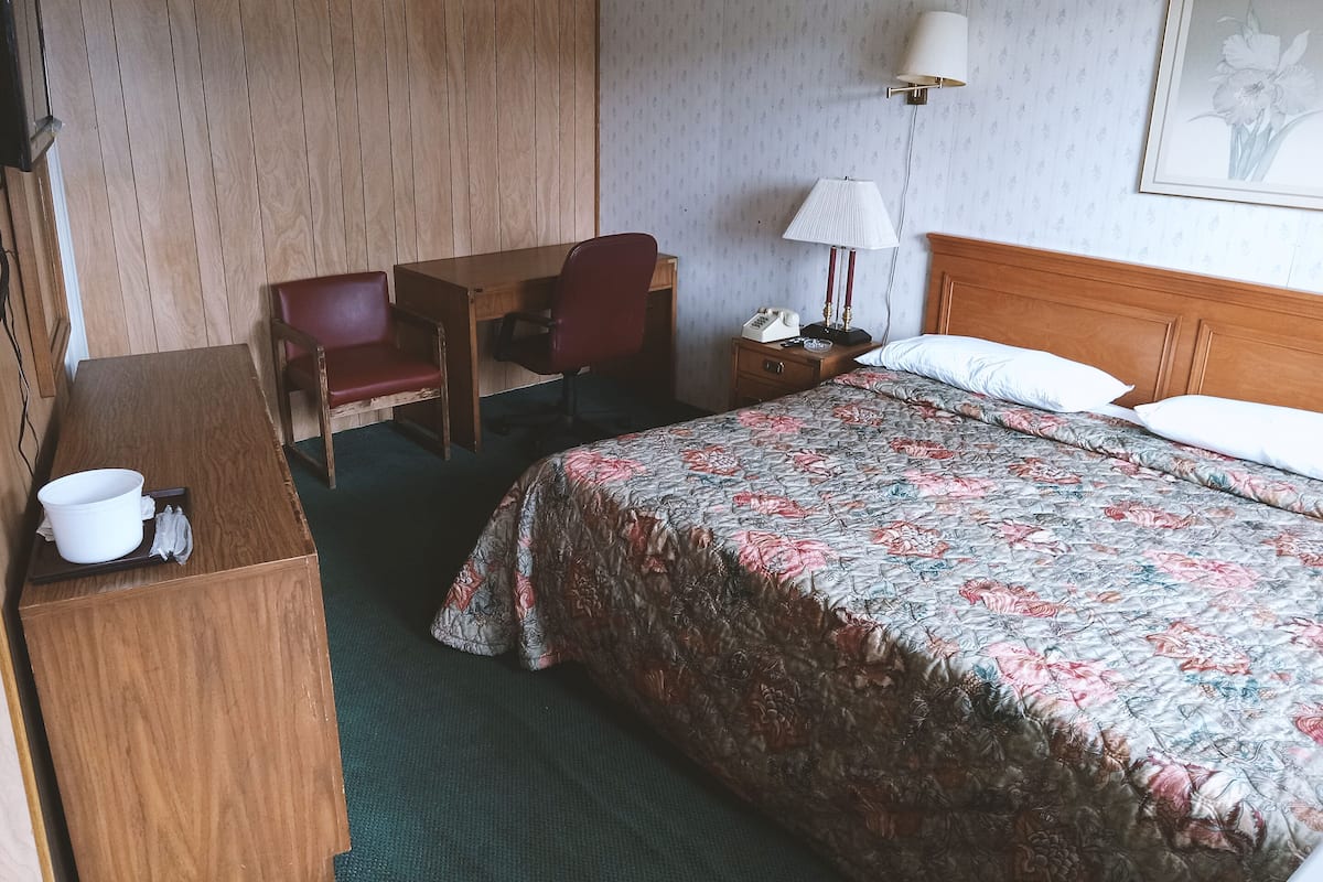 Une chambre d’hôtel avec un bureau en bois, une chaise, un lit avec une literie à motifs floraux, une table de chevet avec une lampe et un tableau encadré au mur.