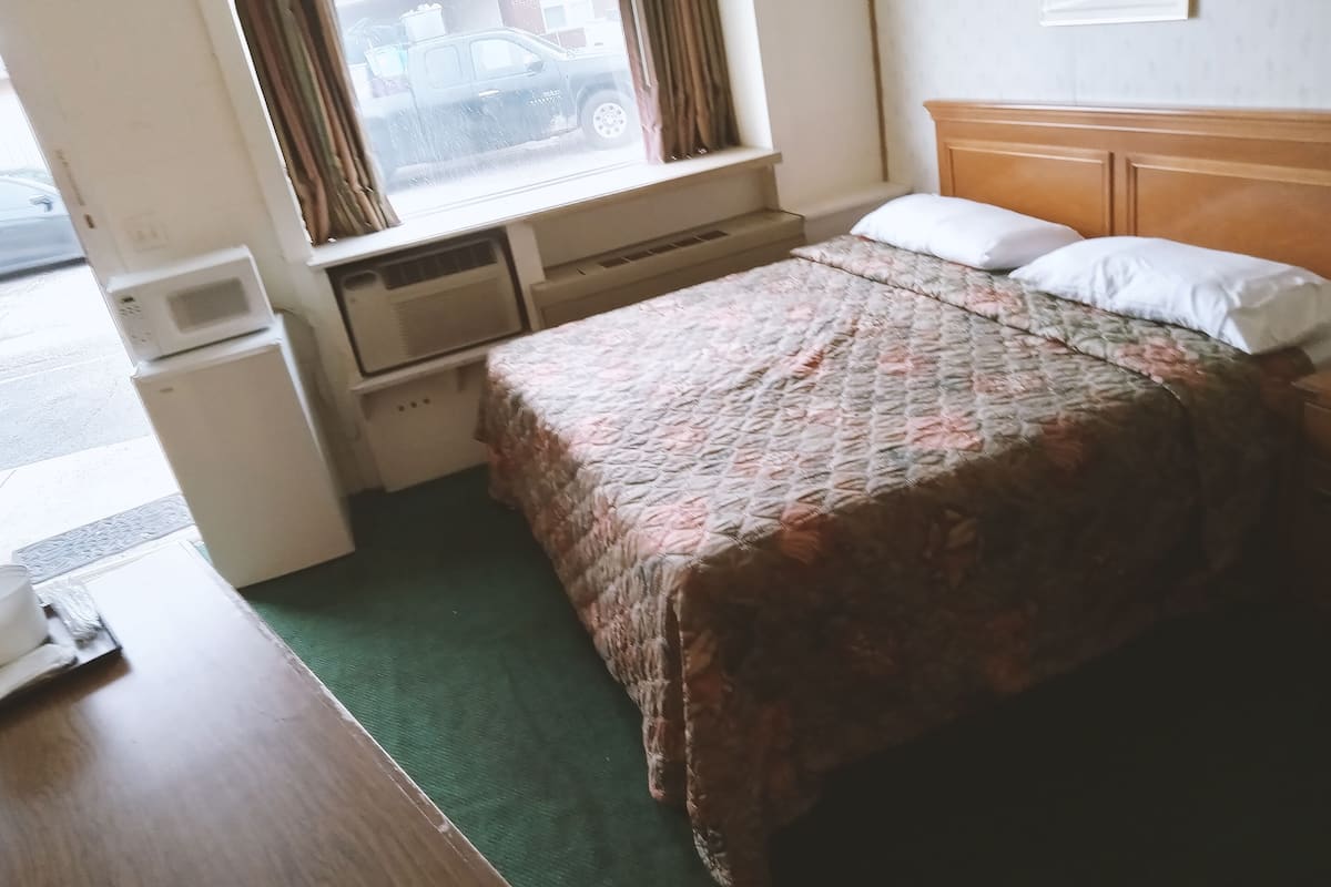 Une petite chambre d’hôtel avec un lit, une tête de lit en bois, une fenêtre avec des rideaux, une moquette verte et une petite table sur laquelle se trouve une tasse.