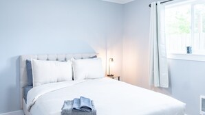 Una cama bien tendida con sábanas blancas y una toalla doblada, una lámpara de noche y una ventana con cortinas.