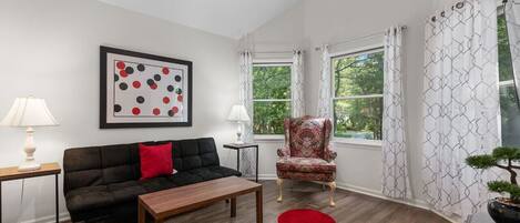 A living room with a black sofa, a wooden coffee table, a red rug, a framed abstract artwork, a patterned armchair, and a window with curtains.