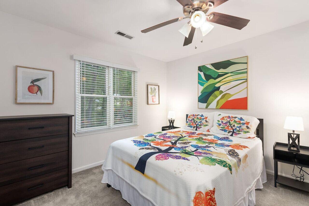 A bedroom with a bed, dresser, ceiling fan, window with blinds, and artwork on the walls.