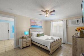 A bedroom with a bed, nightstand, lamp, ceiling fan, and a painting on the wall. - Siesta Key Base | Walk to Beach | Free Activities (Sarasota)