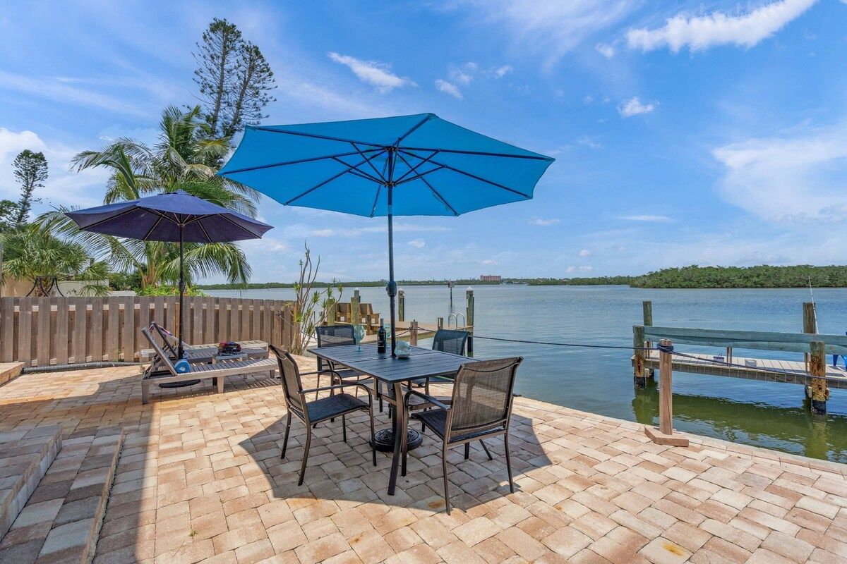 An outdoor patio with a table and chairs, two umbrellas, and a view of a lake.