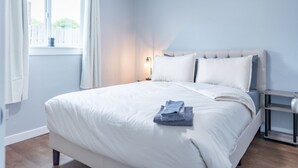 Una cama bien hecha con sábanas blancas y una toalla doblada, una mesita de noche con una lámpara y una ventana con cortinas.