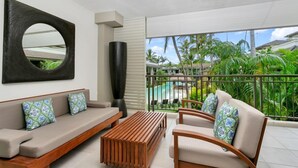 Un salon moderne avec un canapé, un fauteuil et une table basse, une grande peinture abstraite et un balcon offrant une vue sur des palmiers et une piscine.