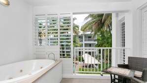Ein modernes Badezimmer mit einer freistehenden Badewanne, einem kleinen Tisch mit einem Sessel und Blick auf einen Pool und Palmen.