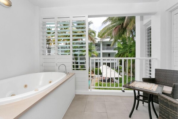 Une salle de bain moderne avec une baignoire sur pieds, une petite table avec une chaise, et une vue sur une piscine et des palmiers.
