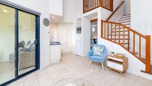 Amplia sala de estar con puerta de cristal que da a un balcón, una escalera de madera y una cocina al fondo.