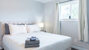 A neatly made bed with a folded towel, a bedside lamp, and a window with curtains. - AquaShores#3 - Pet friendly, BBQ grill & free Wifi (Ocean Shores)