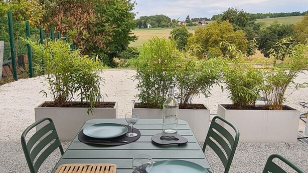 Uma área de refeições ao ar livre com uma mesa posta para dois, rodeada por cadeiras verdes, sob um grande guarda-sol.