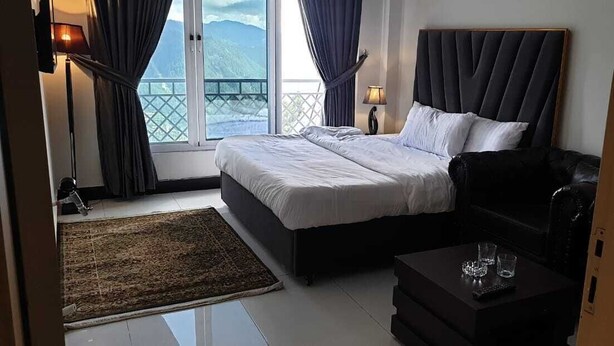 A bedroom with a bed, a black sofa, a small table with glasses, a lamp, and a view of mountains through a large window. - Orangeloft Kalabagh (Abbottabad)