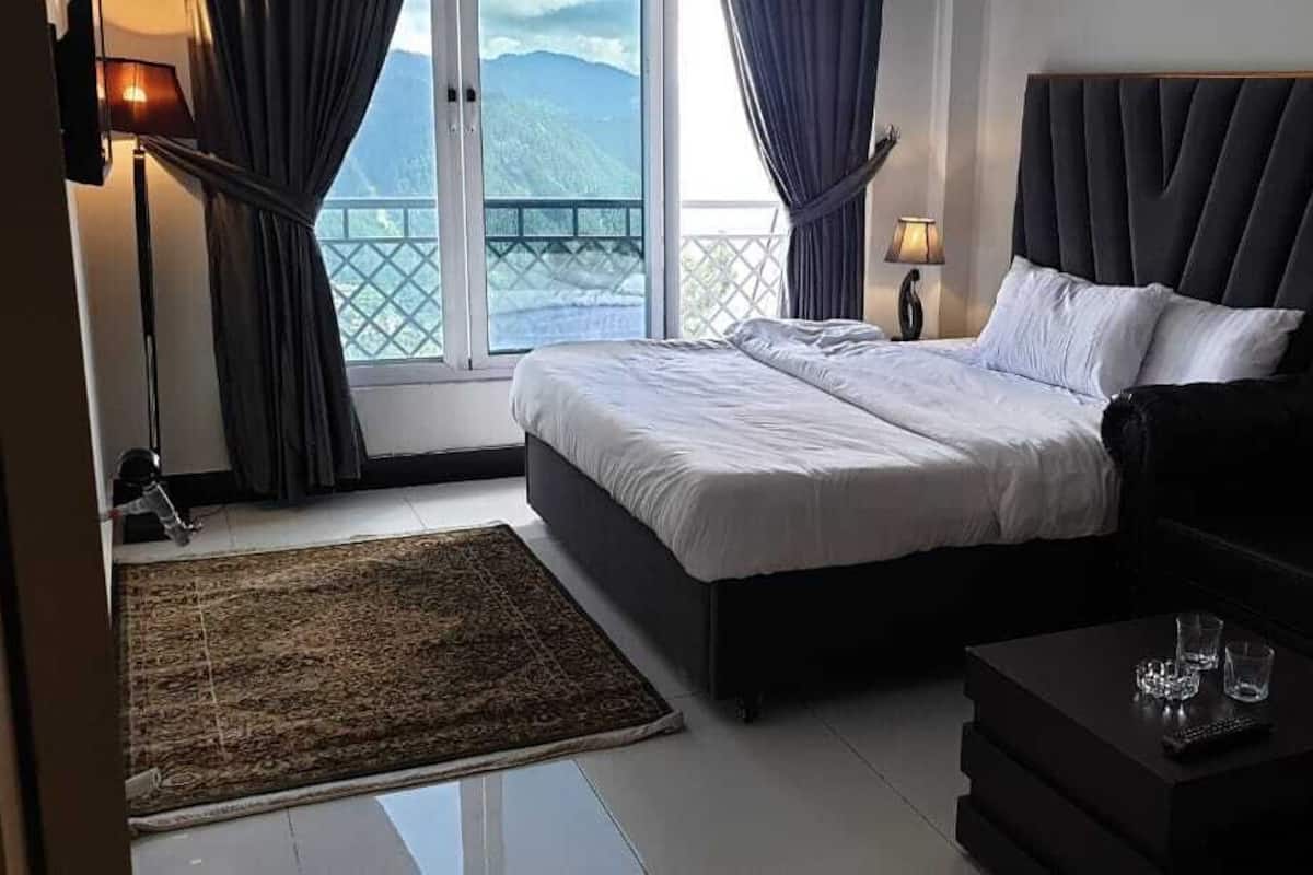 Ein Schlafzimmer mit einem Bett, einer schwarzen Couch, einem kleinen Tisch mit Gläsern, einer Lampe und Blick auf Berge durch ein großes Fenster.