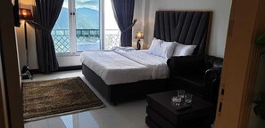 A bedroom with a bed, a black sofa, a small table with glasses, a lamp, and a view of mountains through a large window. - Orangeloft Kalabagh (Abbottabad)