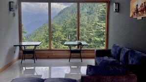 Amplia habitación con un ventanal que ofrece vistas a una montaña boscosa. Hay una mesa negra y un sofá azul con cojines.