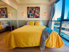 A hotel room with a large bed, a city view, and artwork on the walls.