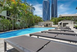 A swimming pool with lounge chairs, palm trees, and modern buildings in the background.
