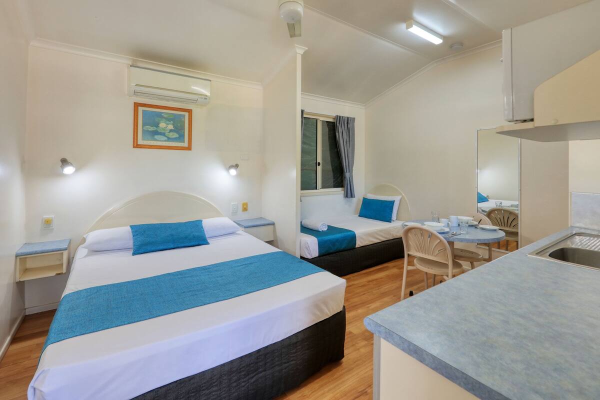 A compact hotel room with two beds, a small kitchenette, and a dining area.
