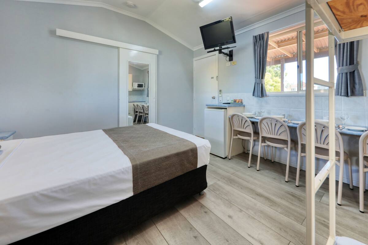 A room with a bed, a desk with chairs, a television, and a kitchenette.