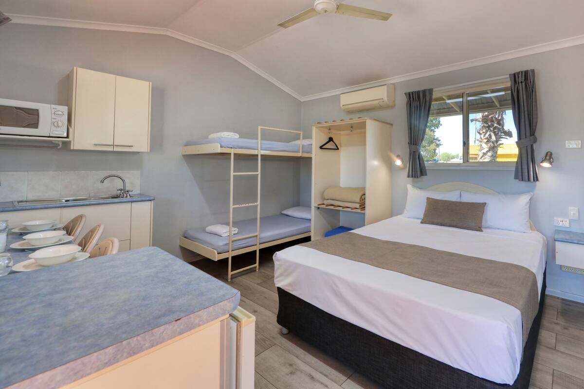 A compact hotel room with a bunk bed, a kitchenette, a single bed, and a window with curtains.
