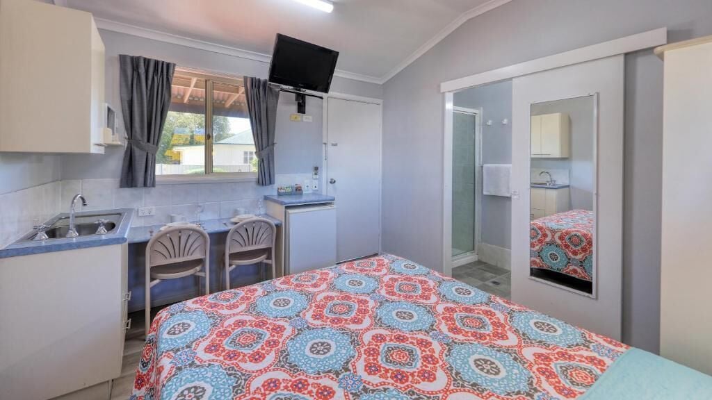 A compact hotel room with a bed, a desk, a chair, a small kitchenette, and a bathroom with a mirror.