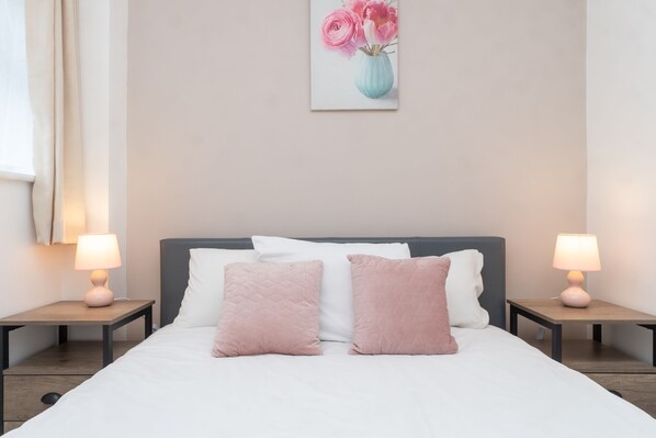 Una cama bien hecha con dos almohadas rosas, una blanca, y un cuadro colgado en la pared de flores rosadas.