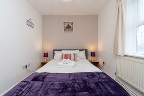 Un lit bien fait, recouvert d’une couverture violette, avec deux lampes de chevet et une œuvre d’art encadrée au-dessus de la tête de lit.