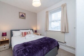 Une chambre à coucher avec un lit, une fenêtre avec des rideaux, une lampe fixée au mur et un radiateur.