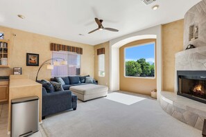 A living room with a fireplace, a ceiling fan, and a large window with a view of trees.