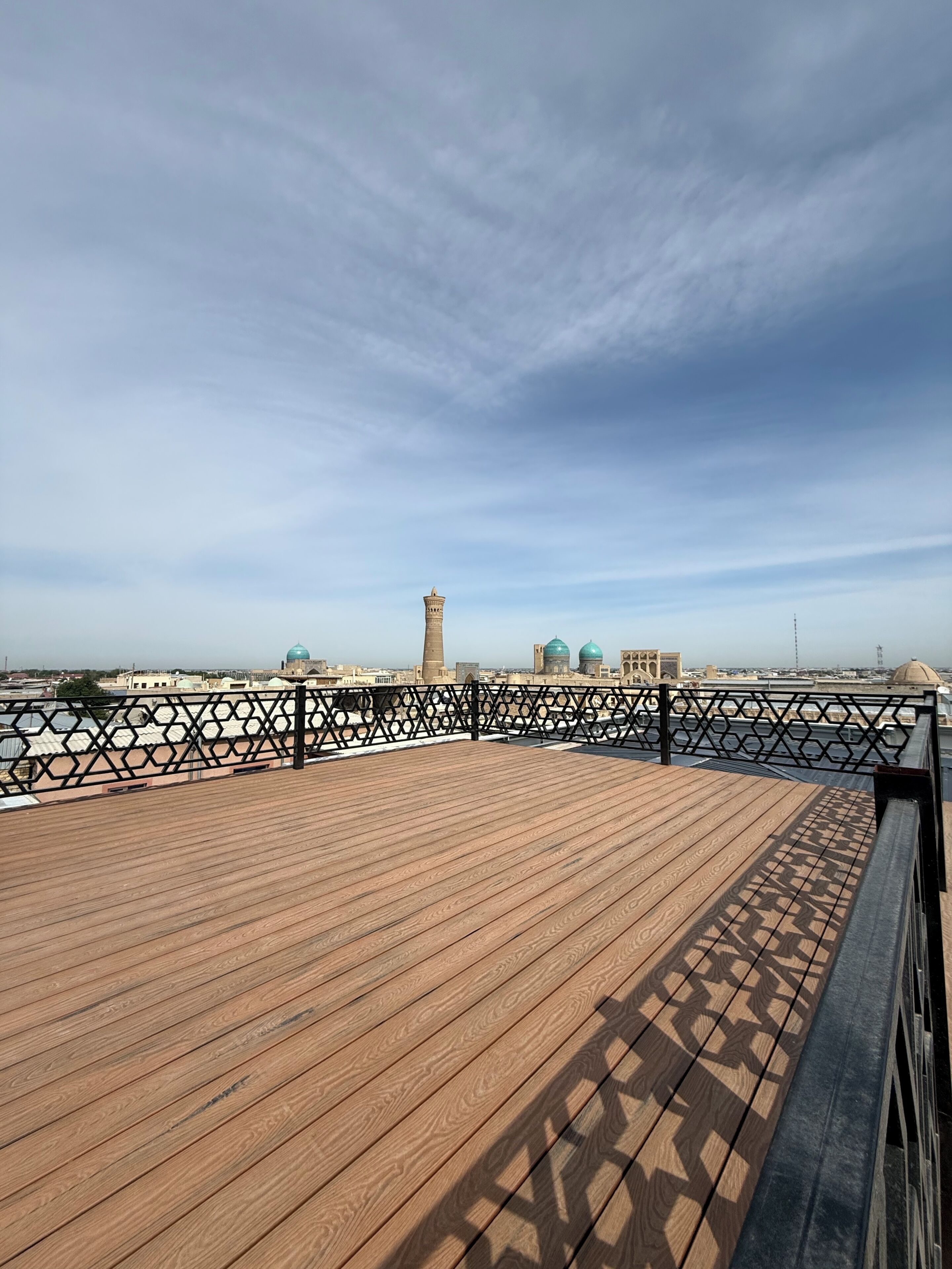 A wooden deck with a metal railing, overlooking a cityscape with buildings and a tower.