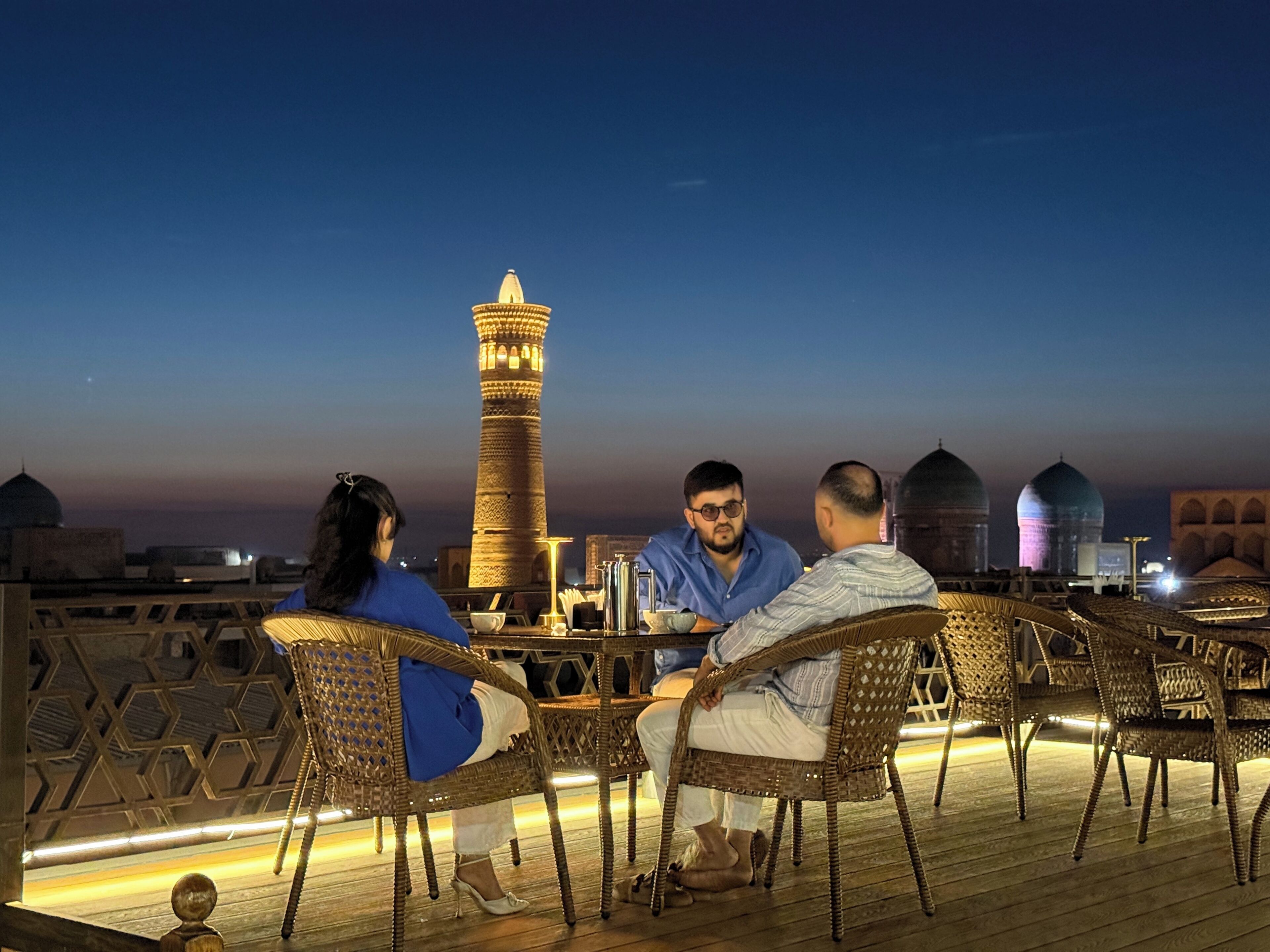 Un groupe de personnes est assis à une table sur une terrasse, d’où l’on peut admirer une haute tour et des bâtiments à coupole.