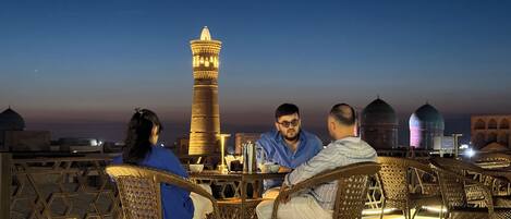 A group of people seated at a table on a deck with a view of a tall tower and domed buildings.